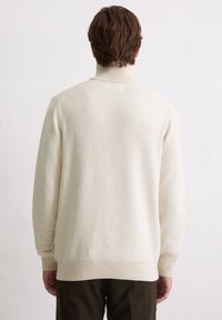 Crème-kleurige trui met opstaande kraag en een textuurafwerking, met ribgebreide boorden en zoom, en een label aan de linkerkant. Eenvoudig ontwerp en zachte stof.