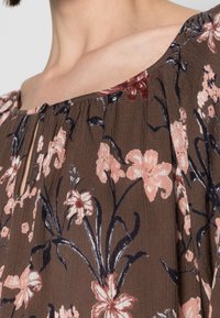 Brun blomsterbluse med korte ærmer, prydet med lyserøde og marineblå blomster, nøglehulsudskæring og lette plisseringer for tekstur.