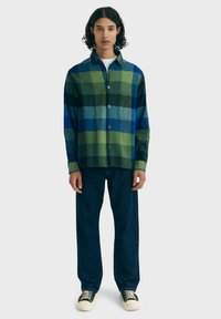 Persoon met krullend haar, gekleed in een groen-blauw geruit overhemd, donkerblauwe jeans en zwart-witte sneakers, staande voor een effen achtergrond.