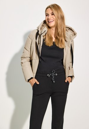 Veste beige rembourrée avec capuche, dotée de cordons de serrage noirs et de poignets élastiques, associée à un pantalon de jogging noir avec un cordon de serrage décoratif.