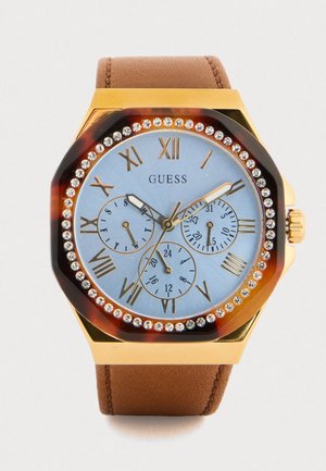 Montre-bracelet Guess ton or avec cadran bleu clair, chiffres romains, trois sous-cadrans, lunette ornée de cristaux et bracelet en cuir marron.