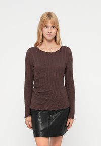 Καφέ τοπ με μακριά μανίκια και υφή με ριμπ σχέδιο. Διαθέτει λαιμόκοψη με κυματιστό περίγραμμα και φοριέται με μαύρη δερμάτινη μίνι φούστα.