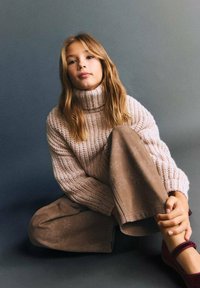 Maglione a collo alto lavorato a maglia di colore rosa chiaro, pantaloni beige larghi e scarpe color bordeaux. La modella ha i capelli lunghi e è seduta in una posa rilassata.