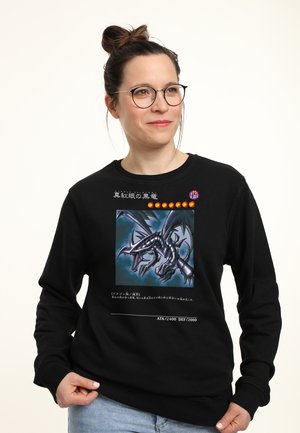 Kvinde med briller iført sort sweatshirt med mørkt dragekunstværk og japansk tekst, stående mod en ensartet lys baggrund.