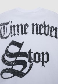 Biała bawełniana koszulka z wyraźnym, czarnym, postarzanym napisem "Czas nigdy się nie zatrzymuje", ozdobnymi elementami i unikalnym krojem pisma.