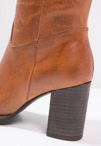 Bottine en cuir marron avec coutures visibles et talon bloc empilé de couleur marron foncé, présentée sur un fond blanc.