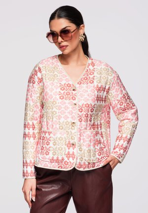 Femme portant de grandes lunettes de soleil teintées de rose, des boucles d’oreilles en or, une veste boutonnée à motif floral rose et beige, et un pantalon brun foncé.