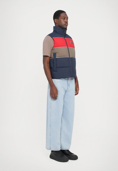 Marineblauwe en rode gewatteerde vest met horizontale kleurblokken, voorzien van een hoge kraag, ritssluiting en zakken aan de voorkant, gedragen over een beige shirt.