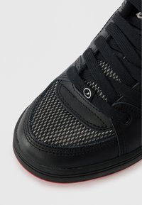 Μαύρο αθλητικό παπούτσι με υφασμάτινο επάνω τμήμα με υφή, δερμάτινες λεπτομέρειες, στρογγυλές θηλιές και κόκκινη καουτσούκ σόλα. Οι κορδόνες είναι περασμένες μέσα από θηλιές υφάσματος.