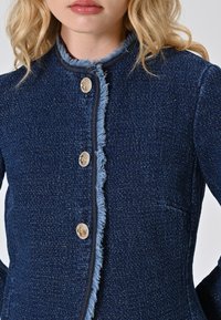 Giovane donna che indossa un giubbotto blu navy con un tessuto strutturato, bottoni dorati e bordi sfilacciati, su uno sfondo neutro.