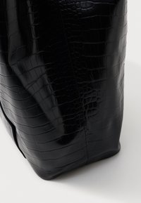 Črna torba iz krokodiljega usnja z embosiranim vzorcem, ki ima gladko teksturo in strukturirano obliko. Ima čiste linije in minimalne šive.