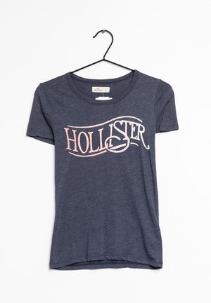 Camiseta de manga corta de color azul marino hecha de tela suave. Presenta un gran gráfico color rosa claro con la palabra "HOLLISTER" impresa en el frente.