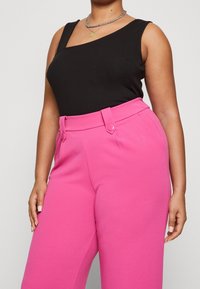 Kvinna som bär en ärmlös svart topp och högmidjade klara rosa byxor med bältesömmar och knappar, som visas från halsen till mitten av låret.