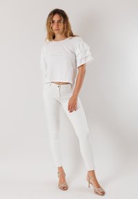 Maglietta bianca corta con maniche corte a volantino, abbinata a jeans bianchi slim-fit e sandali dorati con cinturini. Texture liscia, design minimale.