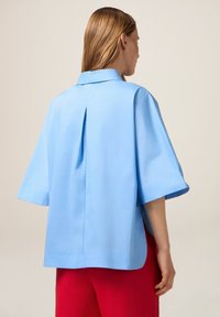 Camicia azzurro chiaro a maniche corte con piega sul retro, realizzata in tessuto morbido. Presenta un colletto classico ed è abbinata a pantaloni larghi rossi.
