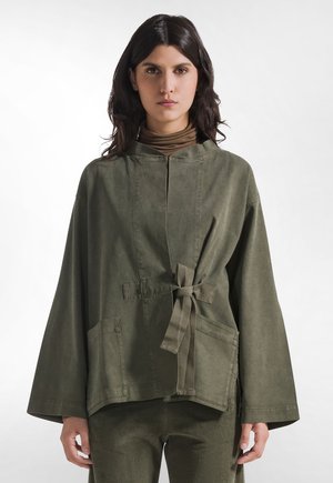 Donna con capelli scuri e mossi indossa una giacca larga verde oliva con laccio frontale e grandi tasche sopra un dolcevita marrone, in piedi davanti a uno sfondo bianco.