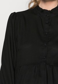 Blusa nera con bottoni e colletto arricciato, dotata di maniche lunghe e design arricciato in vita, realizzata in tessuto morbido.