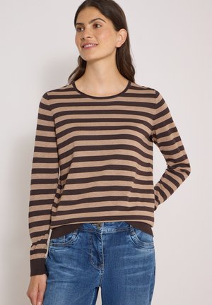 Kvinde iført brun og beige langærmet, vandret stribet skjorte og blå jeans, smilende og kigger let til siden.