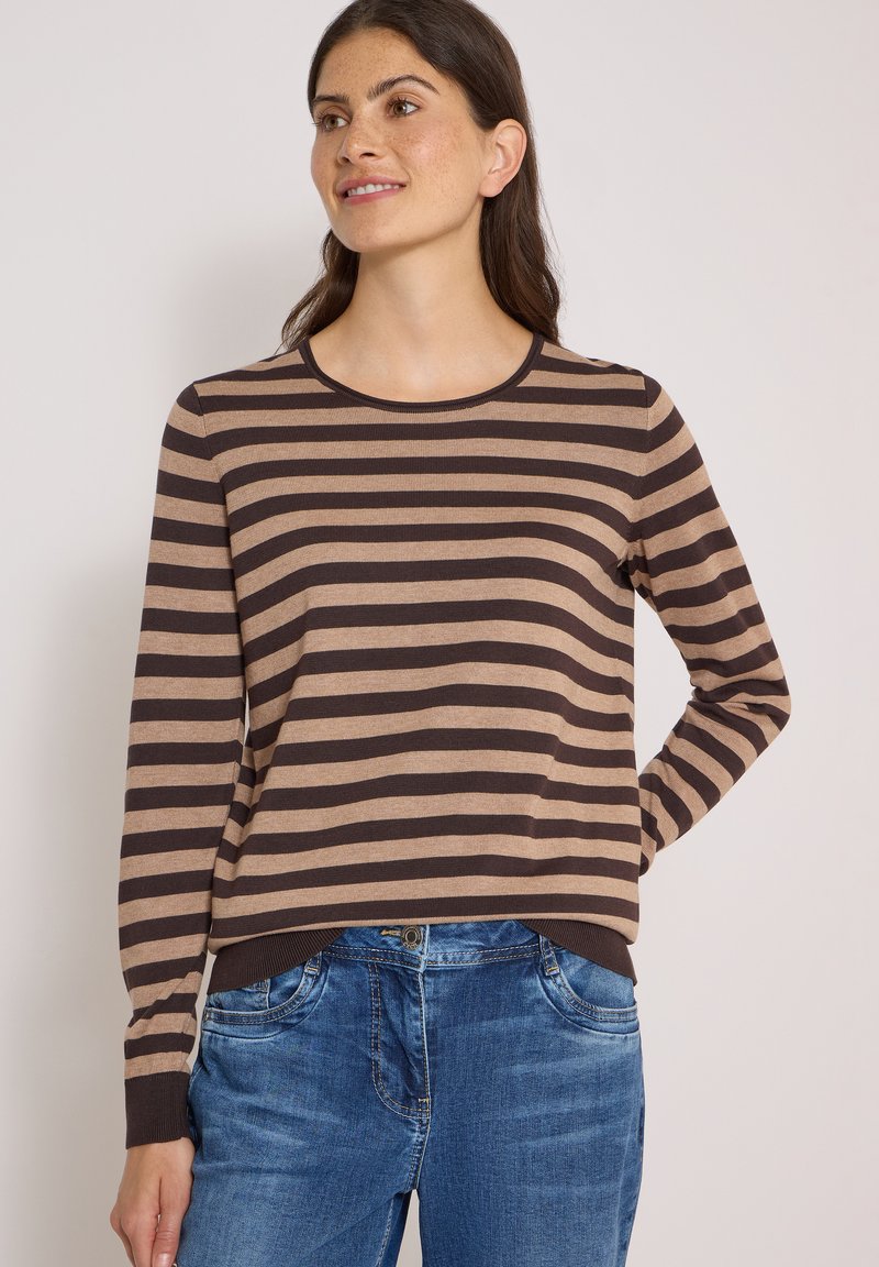 Femme portant un t-shirt à manches longues rayé horizontalement marron et beige et un jean bleu, souriante et regardant légèrement sur le côté.