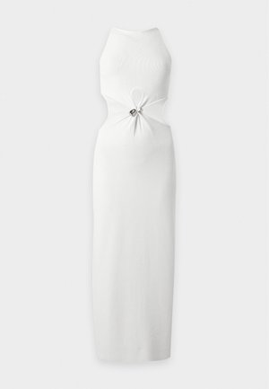 Mujer con vestido blanco sin mangas con aberturas sosteniendo un bolso negro y llevando sandalias negras de tacón alto con tiras sobre un fondo blanco.