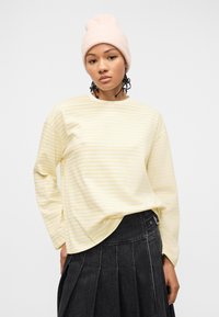 Γυναίκα που φοράει ανοιχτό ροζ πλεκτό σκουφάκι, μακρυμάνικη μπλούζα με κίτρινες και λευκές ρίγες και μαύρη πλισέ φούστα σε απλό φωτεινό φόντο.