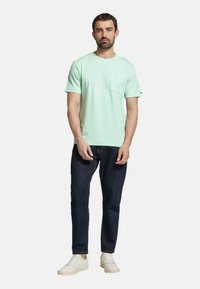 Tricou scurt cu mânecă, de culoare verde mentă, cu un buzunar frontal, asortat cu blugi negri și adidași albi. Textură simplă, croială lejeră.