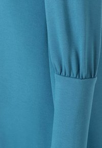 Langärmliges Oberteil in lebhaftem Türrkis, mit texturiertem Stoff und gerafften Manschetten für zusätzliche Details. Glatte Oberfläche mit einem weichen Erscheinungsbild.