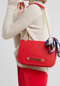 Punainen synteettinen käsilaukku, jossa on kultaketjuolake, etupaneelissa on kohokuvioitu "LOVE MOSCHINO" ja koristeena on koristehuivi.