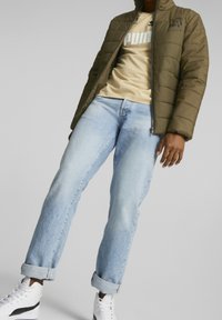Doudoune vert olive, zippée, avec un design matelassé. T-shirt beige avec logo, jean bleu clair, et baskets blanches avec des accents noirs.