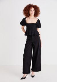 Haut peplum noir texturé avec des manches courtes bouffantes, associé à un jean noir ample et des talons pointus noirs avec des détails décoratifs.