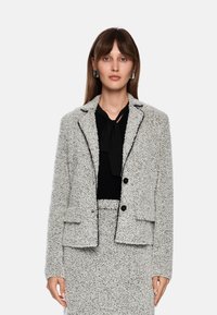 Strukturierter grauer Blazer mit schwarzen Akzenten, Knopfverschluss und zwei vorderen Taschen. Über einem schwarzen Oberteil getragen, das ein Halsband-Detail aufweist.