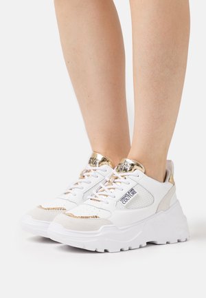 Baskets à plateforme blanches avec des accents dorés et le logo "Versace Jeans Couture" portées sur des jambes nues sur un fond blanc.