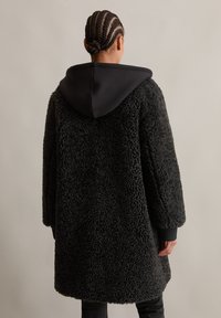 Zwarte faux fur jas met een zachte, gestructureerde afwerking, voorzien van een contrasterende zwarte capuchon en geribbelde manchetten, ontworpen voor een ontspannen pasvorm.