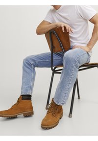 Tan suede lace-up boots with rubber soles, worn with light blue denim jeans. The person is seated on a wooden chair against a neutral background.