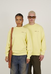 Gule hetter med ribbede mansjetter og halslinjer, med en logotrykk. Modell har på seg en teksturert brun skulderveske sammen med denimjeans.