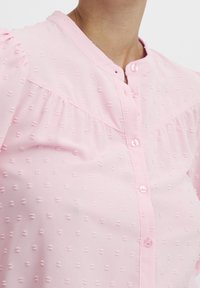 Blusa rosa chiaro con motivo testurizzato, chiusura frontale con bottoni e delicate maniche a palloncino, realizzata in morbido tessuto trasparente.