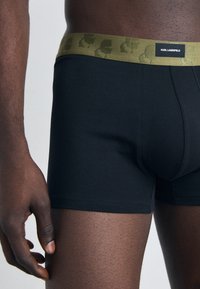 Svarte bomullsboksershorts med en grønn logo i livet som har et gjentakende mønster. Stoffet ser glatt og tettsittende ut.