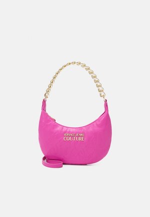 Różowa torebka w kształcie księżyca półksiężyca, z uchwytem z złotego łańcuszka, złotym logo "Versace Jeans Couture" oraz odpinanym paskiem na białym tle.