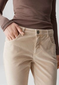 Persoon in beige corduroybroek en een bruin overhemd met lange mouwen, met de linkerman rustend in de broekzak.