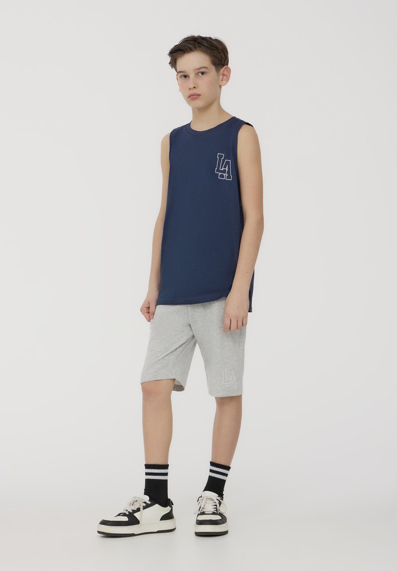 Canottiera senza maniche blu marina con logo bianco "LA", abbinata a pantaloni corti grigi chiari con logo coordinato; indossata con sneakers bianche e nere.