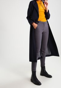 Lungo cappotto blu su un maglione a coste senape, abbinato a pantaloni grigi aderenti e stivali neri con lacci. Texture morbide e vestibilità su misura.