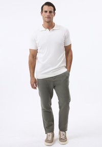 Weißes Polohemd mit kurzen Ärmeln und klassischem Kragen, kombiniert mit hell olivgrünen Hosen und beigen Sneakers mit weißen Sohlen.