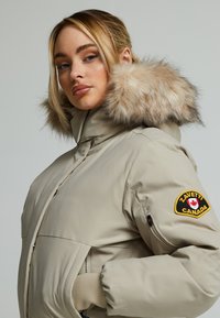 Beige péřová bunda s umělou kožešinou na kapuci, předním zipem, elastickými manžetami, bočními kapsami a logem na rukávu. Hladký, voděodolný materiál.