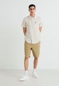 Lysestripete kortermet skjorte, beige shorts og hvite sneakers. Har en brystlomme og en avslappet passform. Glatt stofftekstur.