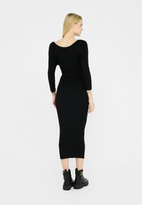 Una donna vista di spalle indossa un vestito nero aderente con maniche lunghe, abbinato a stivaletti neri, su uno sfondo bianco semplice.