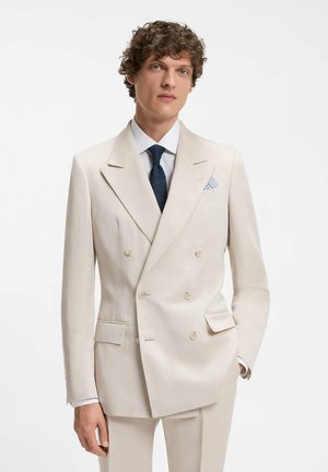 Hombre vestido con un traje cruzado crema, camisa blanca, corbata texturizada azul marino y pañuelo azul claro en el bolsillo, sobre un fondo blanco.