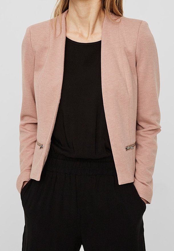 Femme portant un blazer ajusté rose clair par-dessus un haut noir et un pantalon noir à taille élastique, les mains dans les poches, debout devant un fond uni.