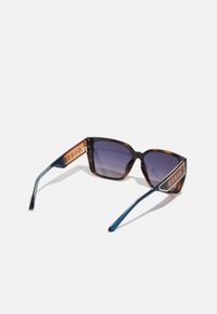 Occhiali da sole in tartaruga con lenti scure a gradiente, forma rettangolare e aste blu a contrasto con un accento del logo dorato "GUESS".