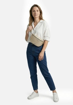 Kobieta w białej koszuli, niebieskich dżinsach i białych sneakersach, nosząca beżową torbę na ramię, stojąca na tle jednolitego białego tła.