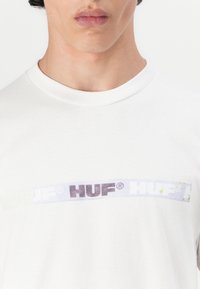 Bijela pamučna majica s horizontalnim grafičkim prikazom na kojem se nalazi natpis "HUF" u raznim bojama i teksturama, uključujući ljubičaste i zelene naglaske.